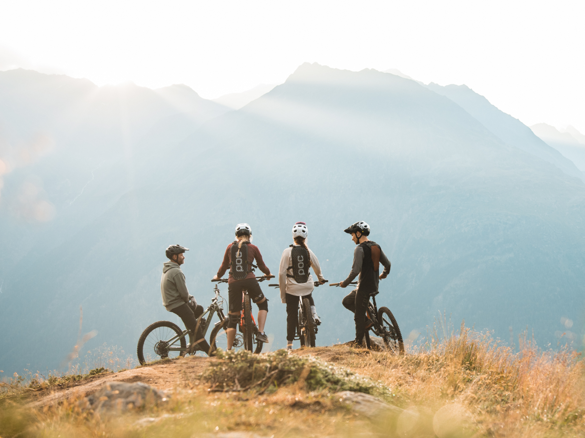 Vier Radfahrer stehen mit Fahrrädern auf einem Hügel und blicken in die Berge bei Sonnenaufgang, vor alpiner Kulisse.