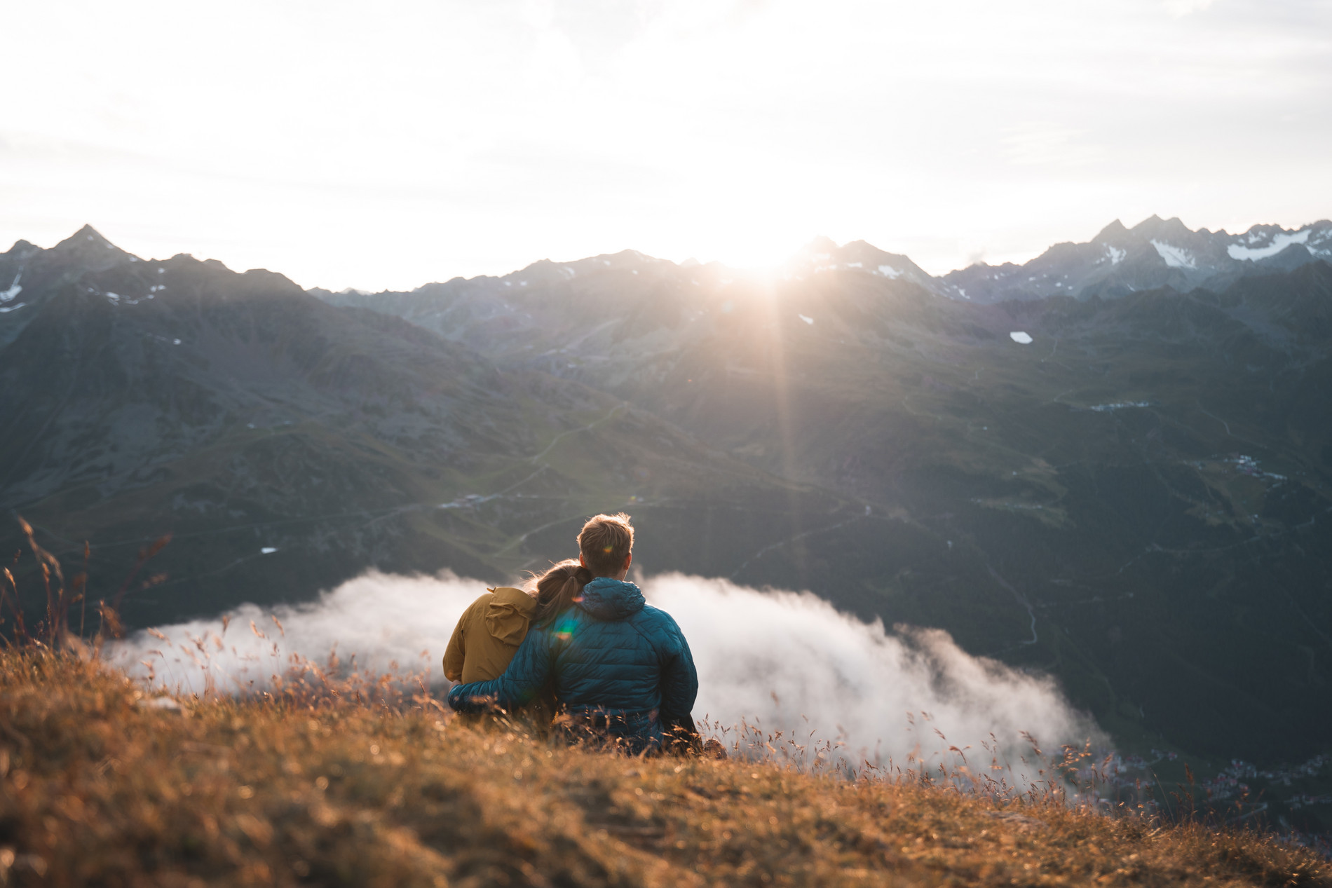 Ein Paar sitzt auf einem Hügel und schaut in einen Sonnenuntergang über einer bergigen Landschaft.