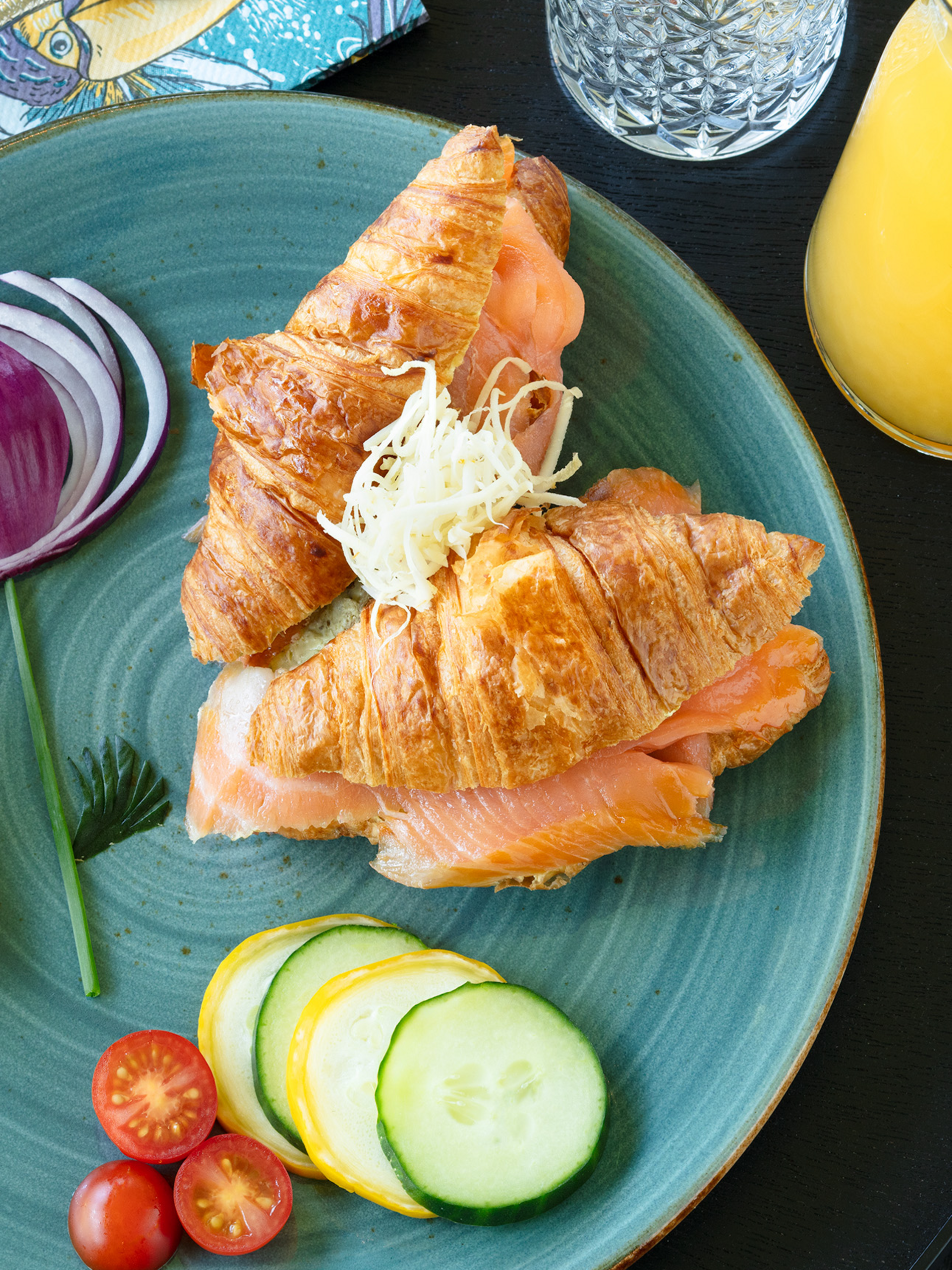 Croissant gefüllt mit Lachs auf blauem Teller, neben Tomaten, Zwiebel, Zitrone und Gurkenscheiben, mit Saft und Besteck.