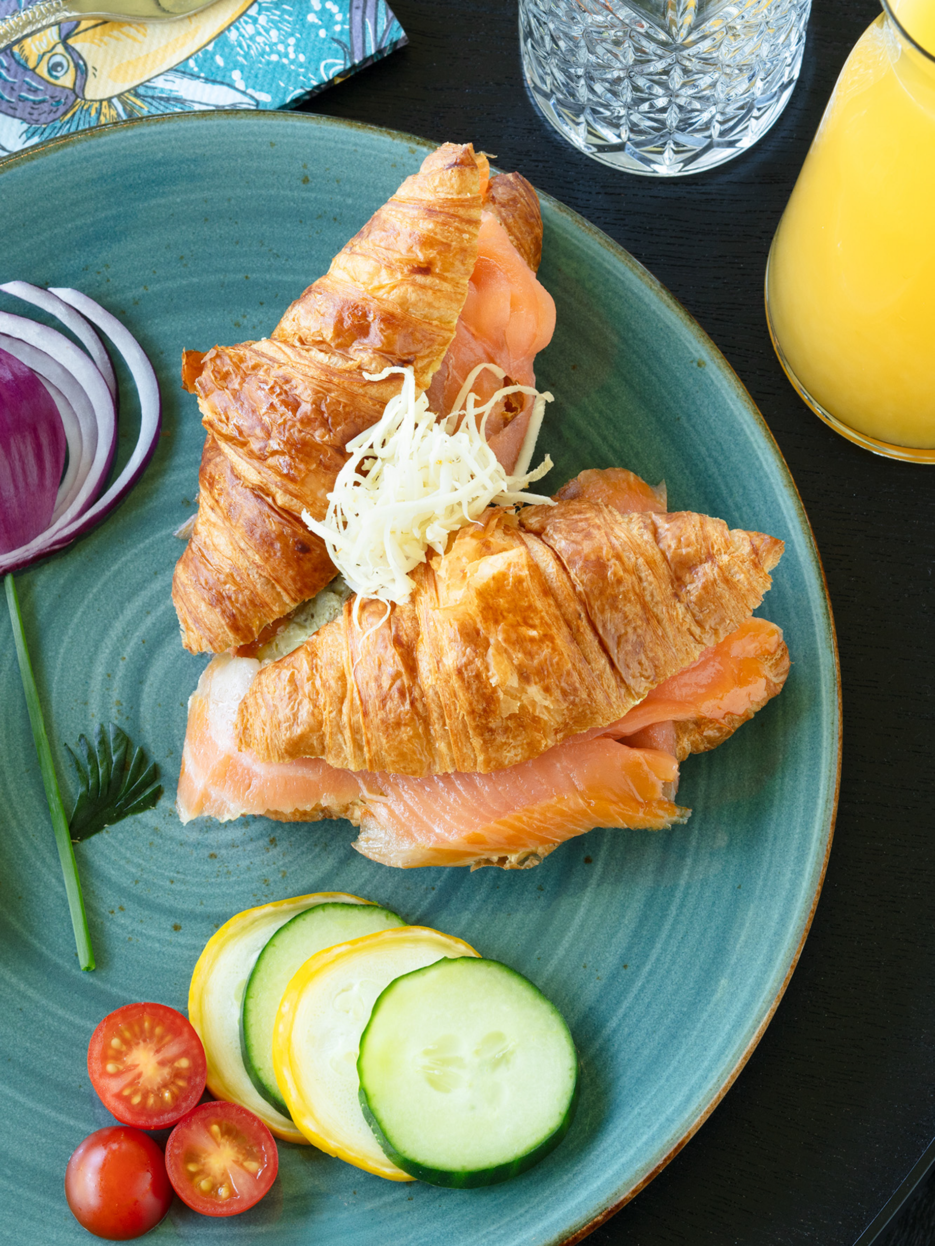 Croissant gefüllt mit Lachs auf blauem Teller, neben Tomaten, Zwiebel, Zitrone und Gurkenscheiben, mit Saft und Besteck.