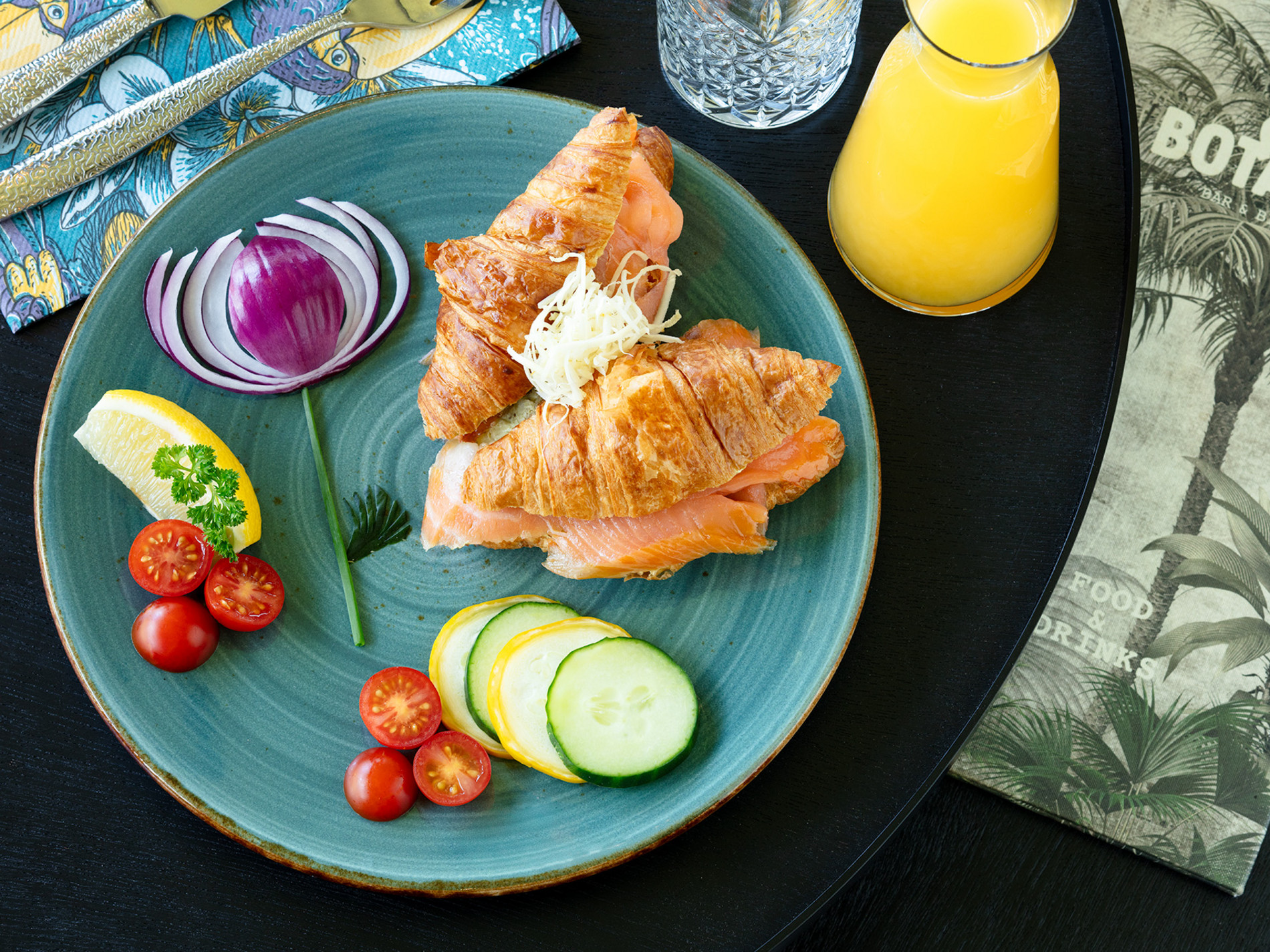 Croissant gefüllt mit Lachs auf blauem Teller, neben Tomaten, Zwiebel, Zitrone und Gurkenscheiben, mit Saft und Besteck.