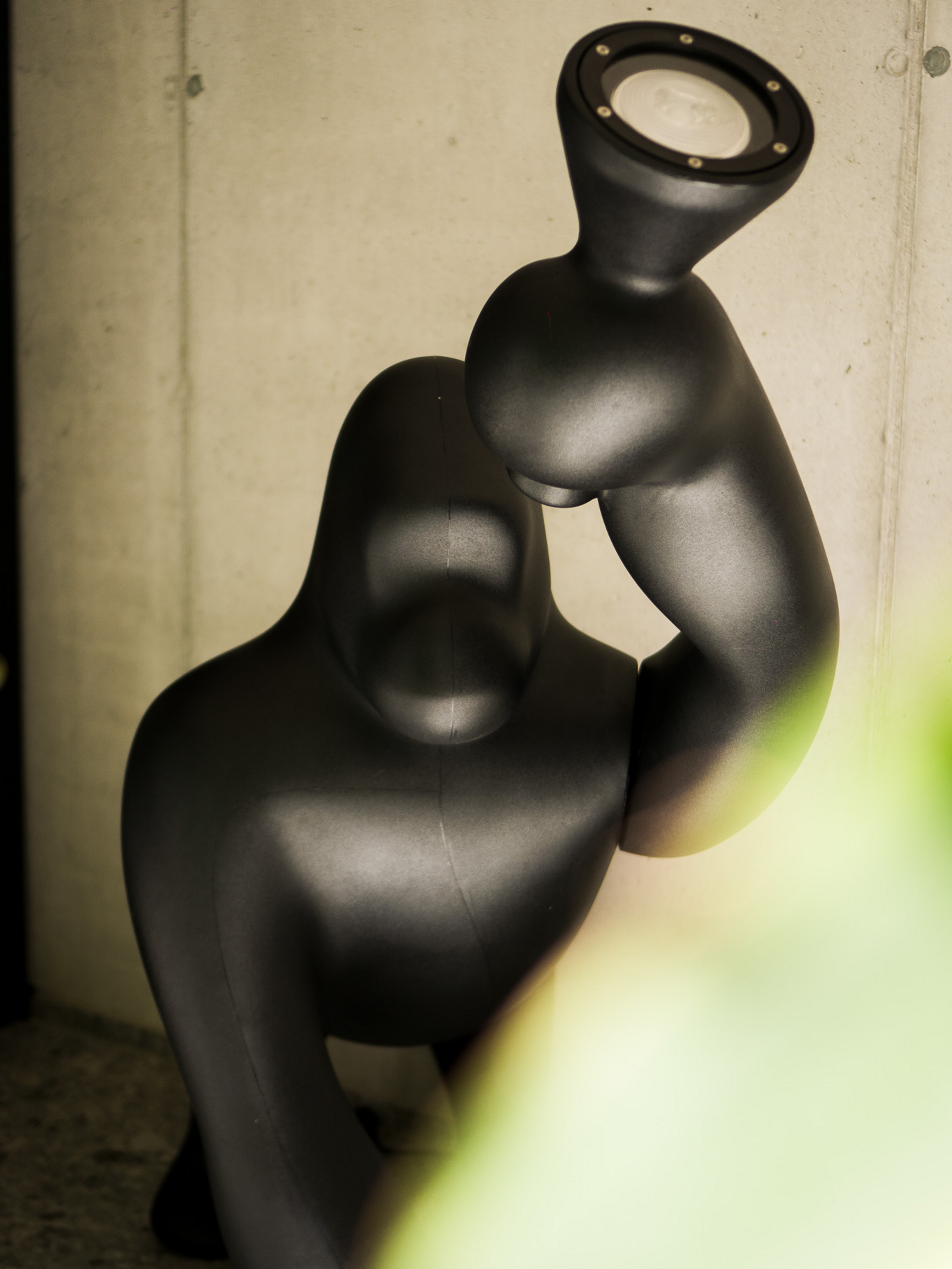 Eine schwarze Skulptur hält einen Leuchtstrahler hoch, platziert vor einer Betonwand mit unscharfem Grün im Vordergrund.