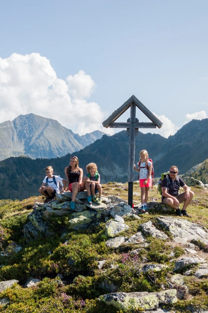 Personen sitzen und stehen auf einer Bergwiese neben einem Holzkreuz im alpinen Bergpanorama bei klarem Himmel.