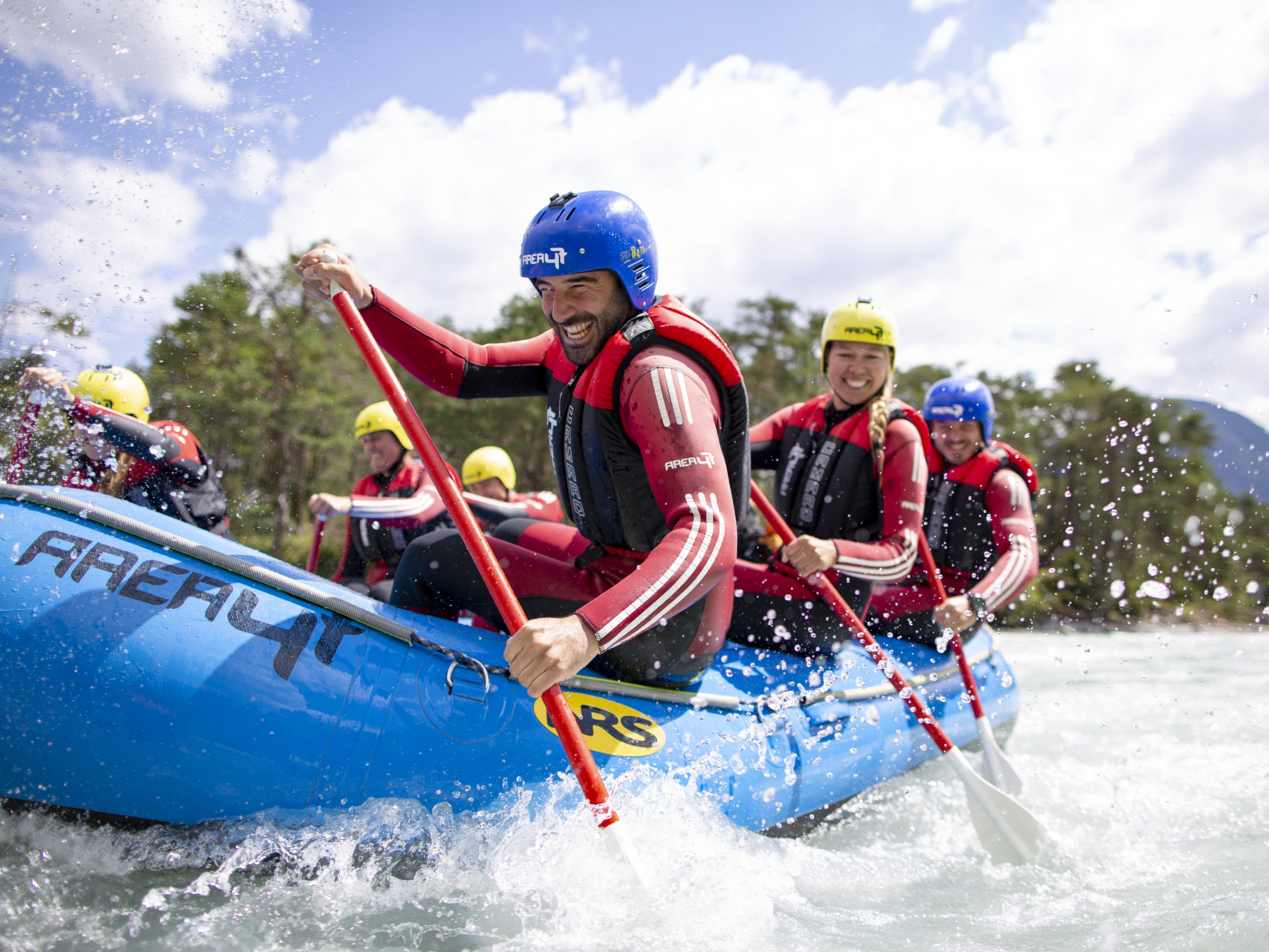 Personen paddeln ein Schlauchboot in Fluss, tragen Helme und Schwimmwesten, Wasser spritzt, Berge und Bäume im Hintergrund.
