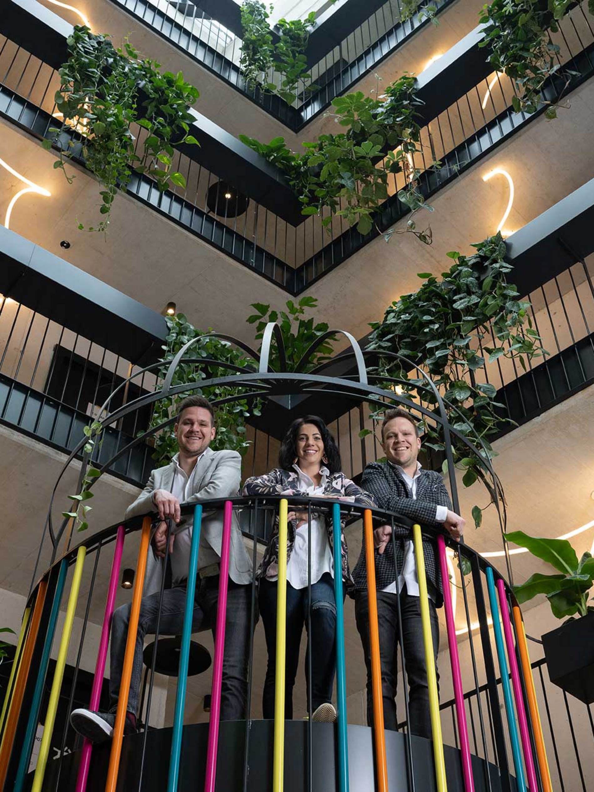Drei Personen lehnen auf einem bunten Geländer, umgeben von Pflanzen in einem lichterfüllten Atrium mit mehreren Etagen.