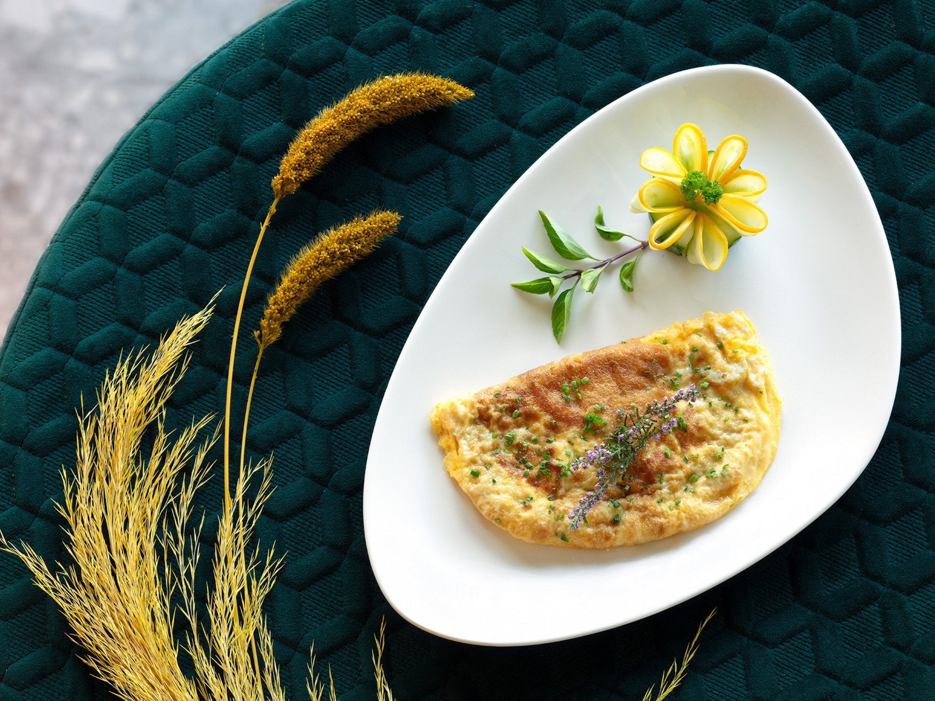 Omelett dekorativ mit Kräutern und Blume auf weißem Teller, arrangiert auf grüner, gemusterter Tischdecke mit Gräsern.