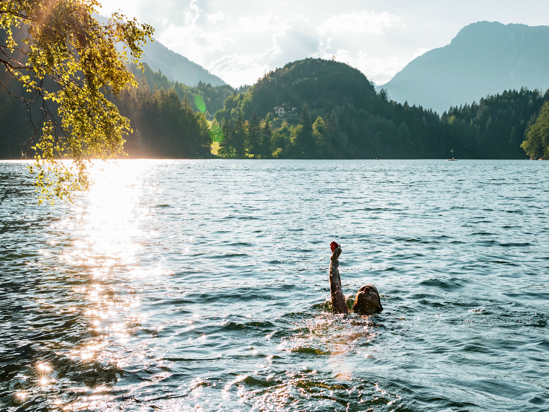 Person schwimmt im See, hebt Arm, umgeben von Bergen und Bäumen, Sonnenlicht reflektiert im Wasser, vor alpiner Kulisse.
