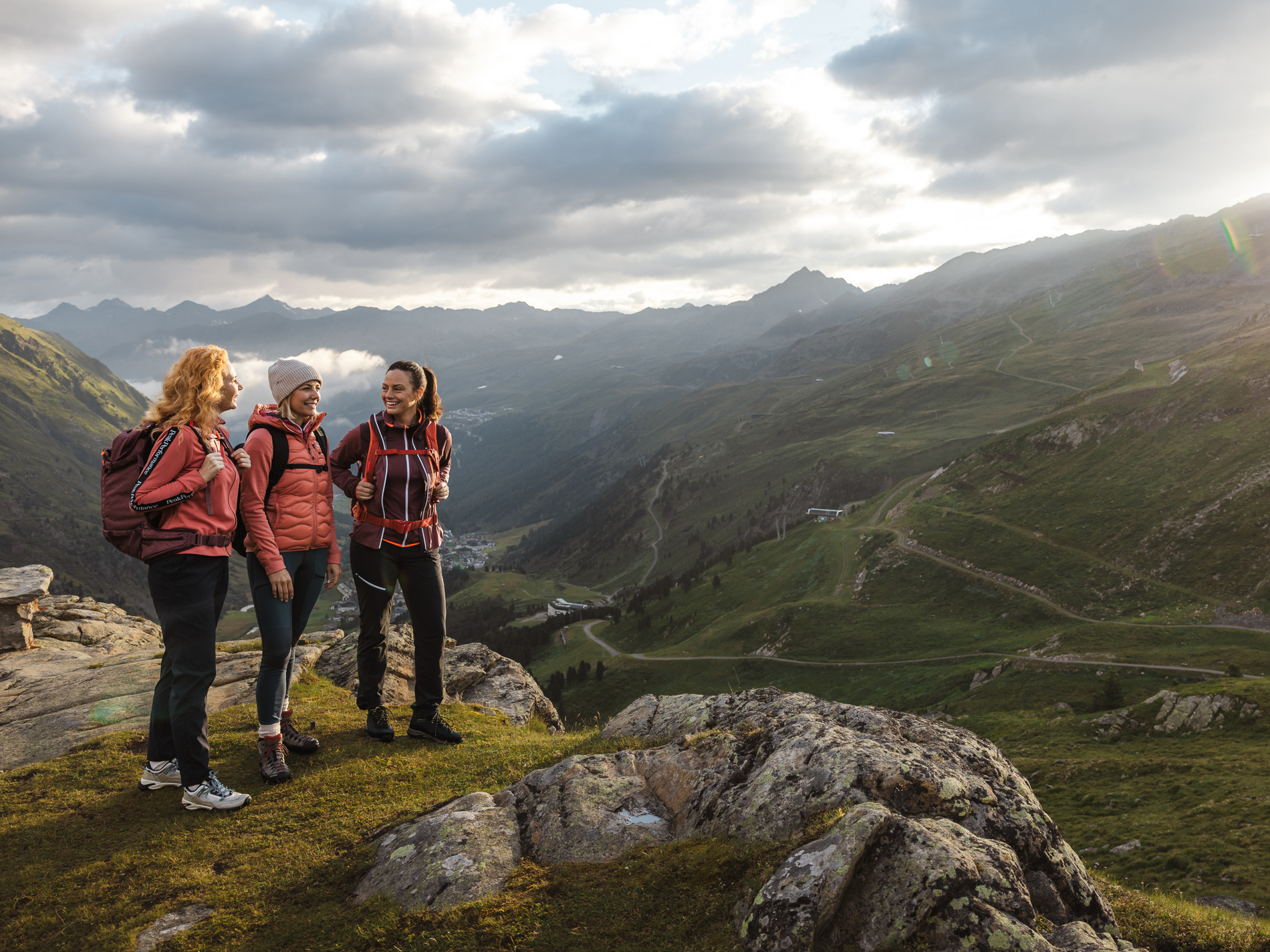 Drei Personen stehen lächelnd auf einem Bergpfad mit Rucksäcken, bei Sonnenaufgang, umgeben von einer Berglandschaft.
