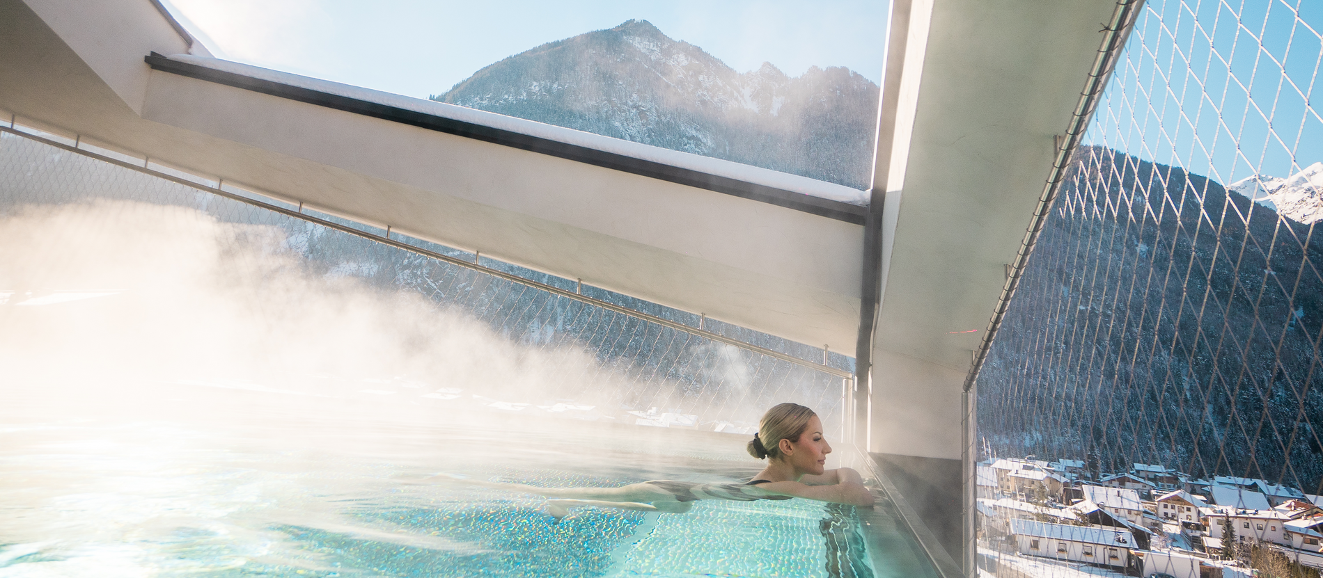JUNGLE VIBES & ALPENLUFT Eine Frau entspannt im dampfenden Pool auf einem Balkon, mit schneebedeckten Bergen im Hintergrund, vor alpiner Kulisse.