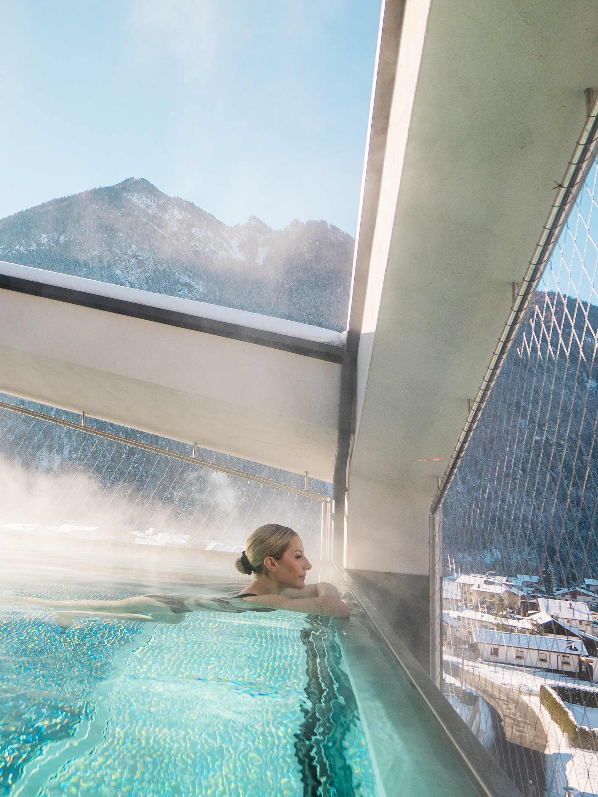 Eine Frau entspannt im dampfenden Pool auf einem Balkon, mit schneebedeckten Bergen im Hintergrund, vor alpiner Kulisse.