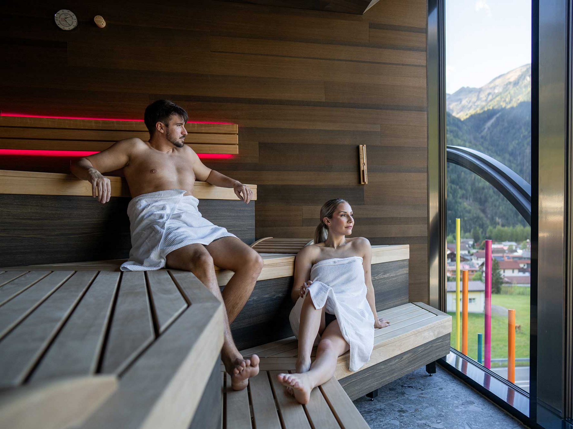 Zwei Personen sitzen entspannt in einer Sauna, umgeben von hölzernen Bänken und Wänden. Sie tragen weiße Handtücher.