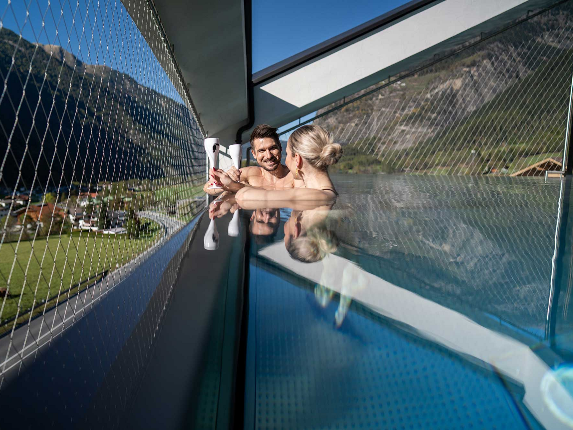 Zwei Personen entspannen sich im Infinity-Pool auf einer Terrasse mit Blick auf Berge und blauen Himmel im Hintergrund.