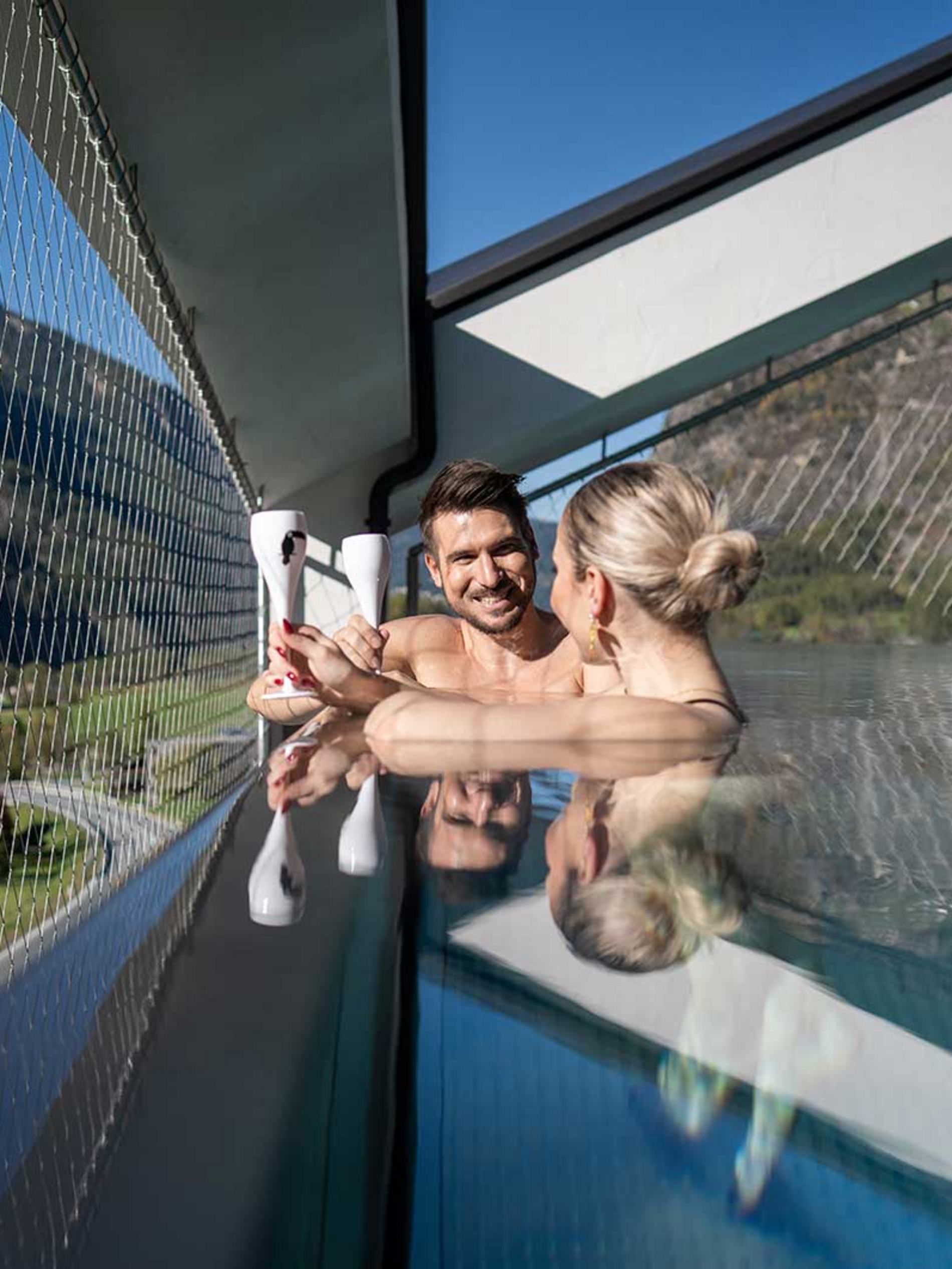 Zwei Personen entspannen sich im Infinity-Pool auf einer Terrasse mit Blick auf Berge und blauen Himmel im Hintergrund.