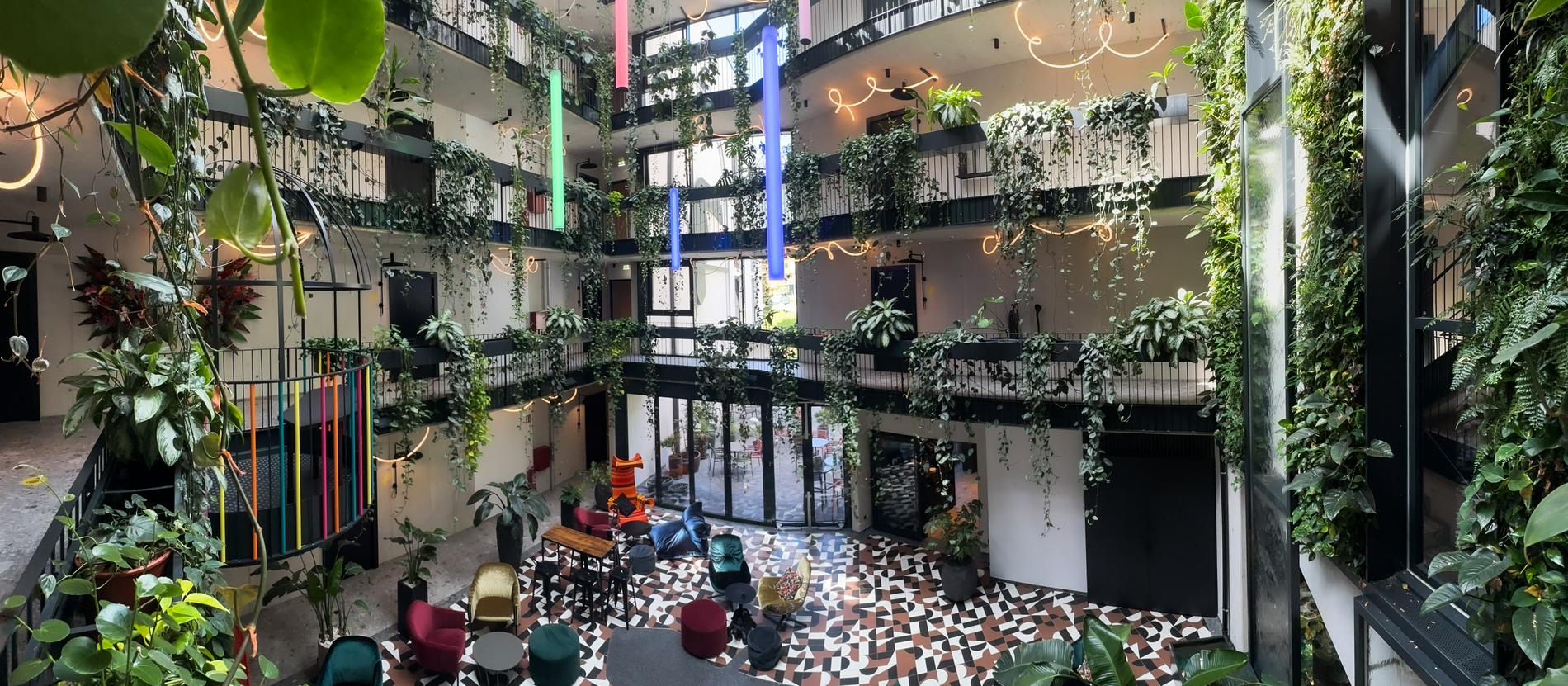 JUNGLE VIBES & ALPENLUFT Grüner überdachter Innenhof mit Pflanzen, bunten Leuchten und modernem Mobiliar umgeben von mehreren Etagen mit Balkonen.