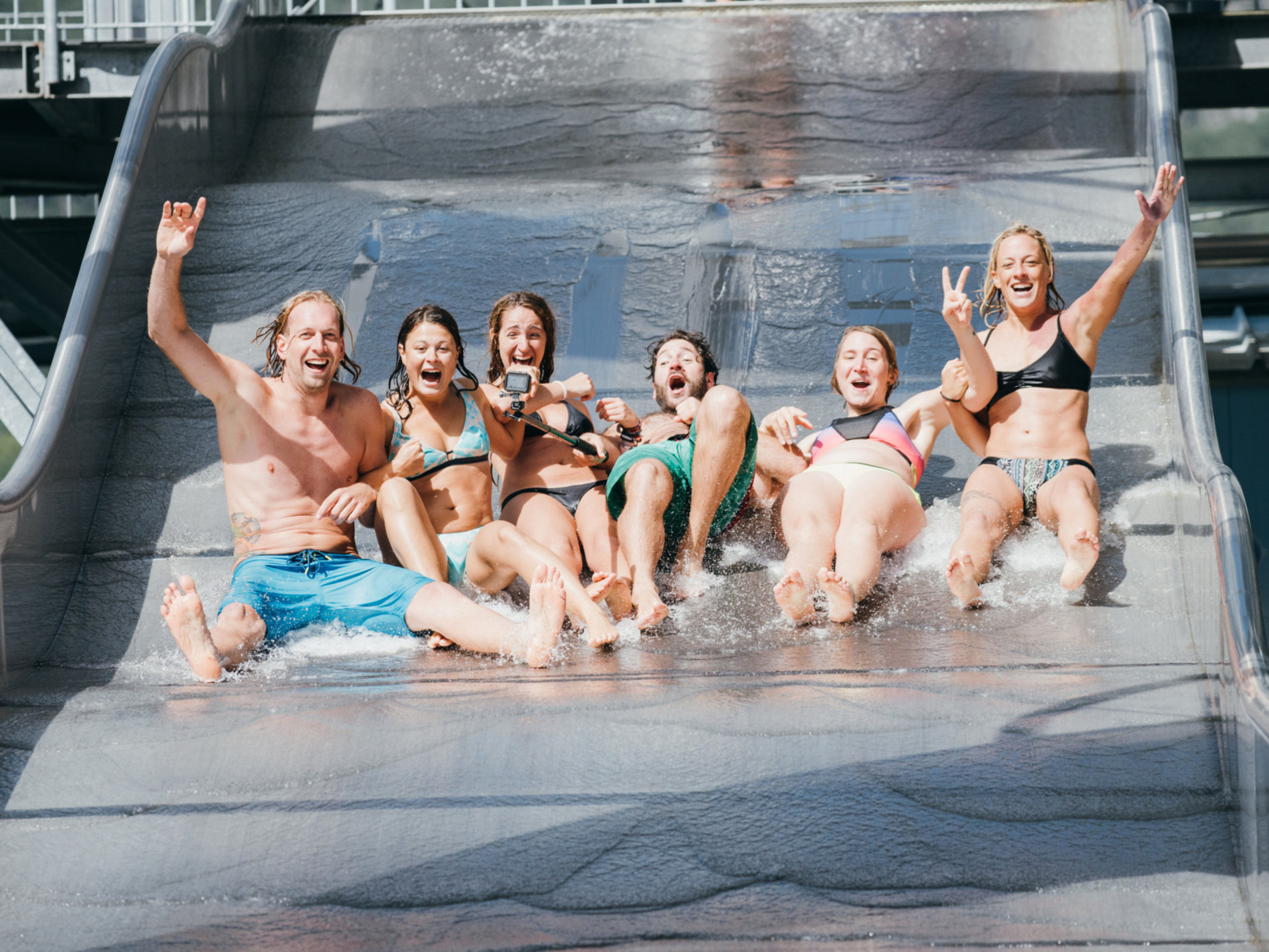 Gruppe Menschen rutscht lachend nebeneinander eine Wasserrutsche hinunter.