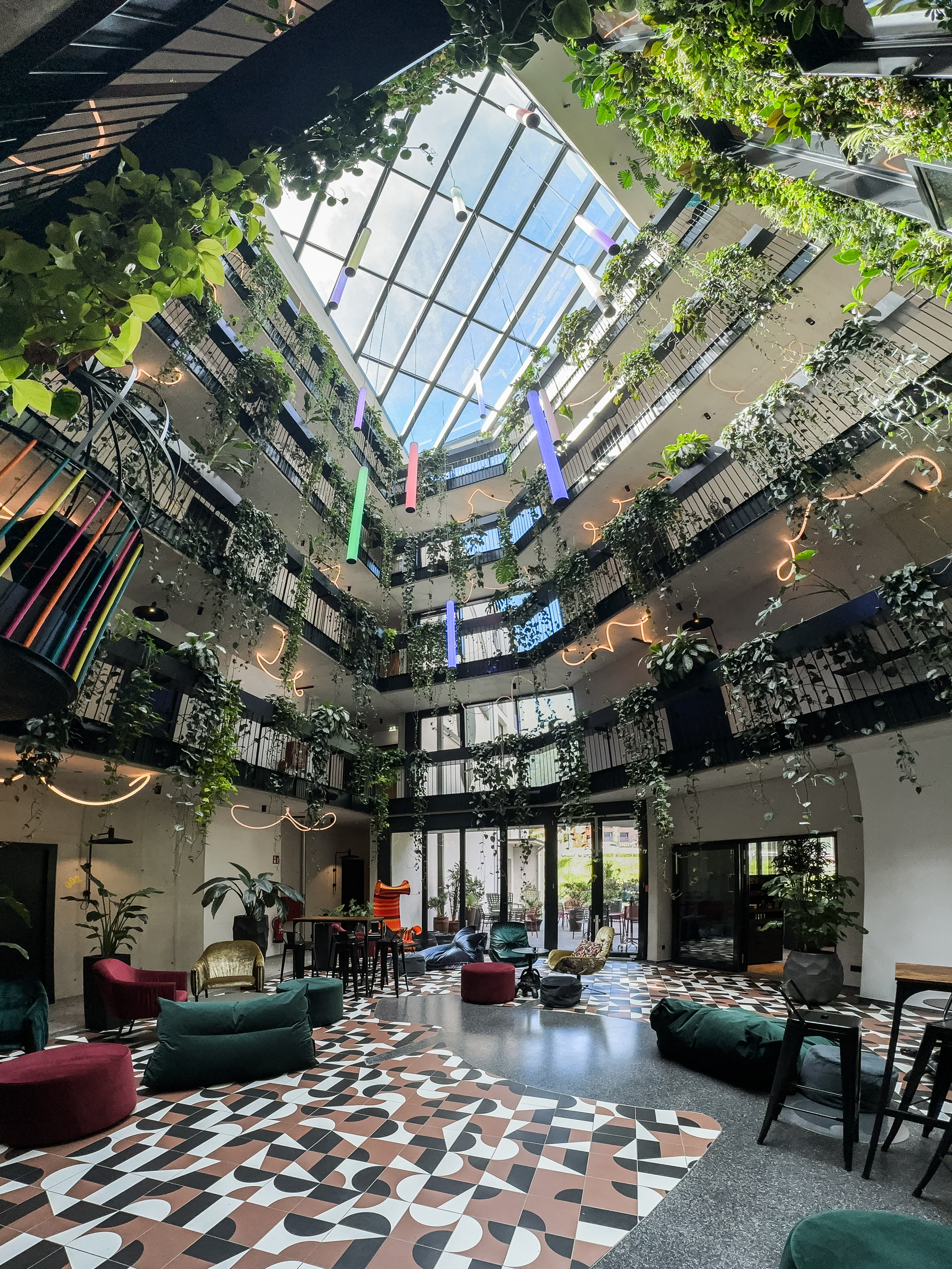 Sofas und Stühle stehen in hellen, begrünten und überdachten Innenhof mit hängenden Pflanzen, farbigen Elementen und Boden.