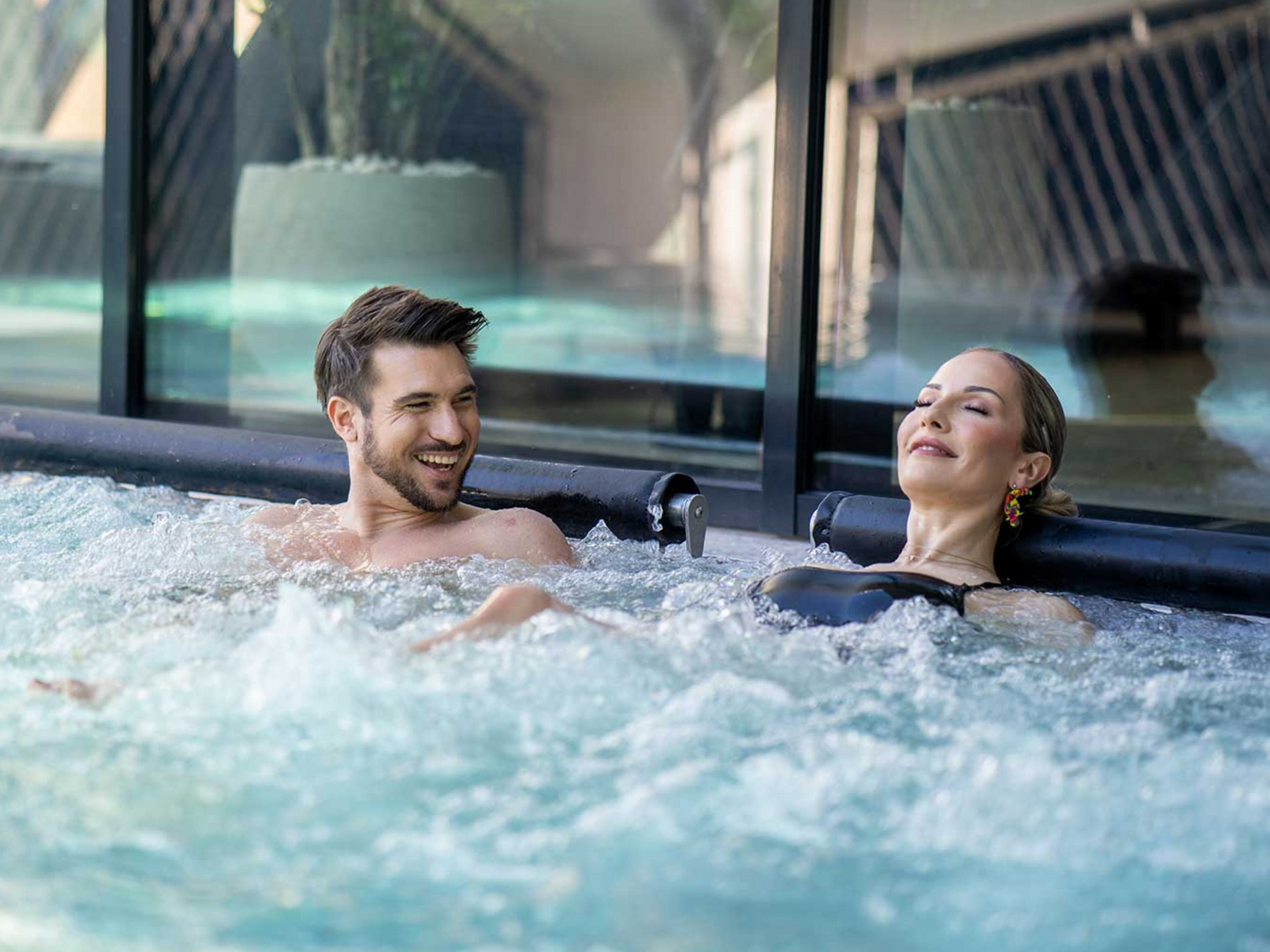 Zwei Personen entspannen im sprudelnden Whirlpool neben großen Fenstern und genießen das Wasser in einem modernen Spa.