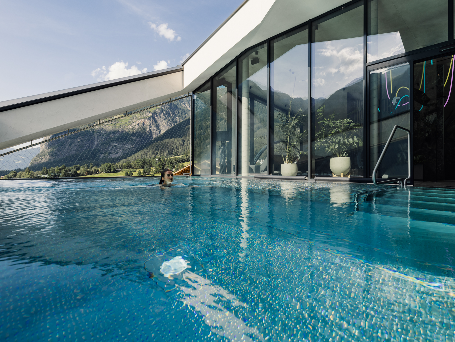 Person schwimmt im Pool vor modernem Glasgebäude, im Hintergrund sind Berge und blauer Himmel zu sehen.